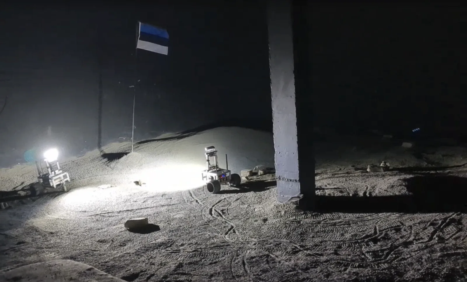 Two rovers driving on Moon-like sand inside a dark analogue hall, lit by their own headlights